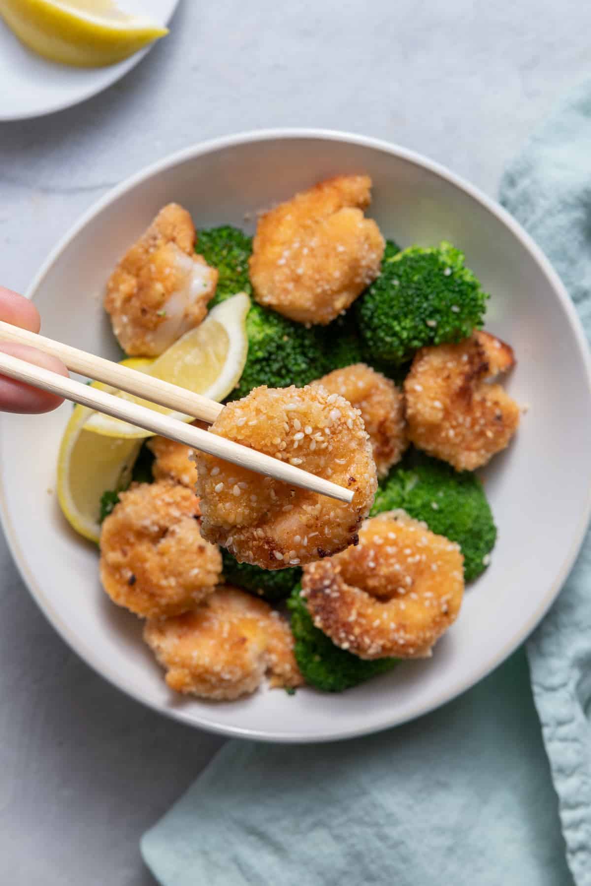 Chopstick holding sesame shrimp over plate of the shrimp with broccoli