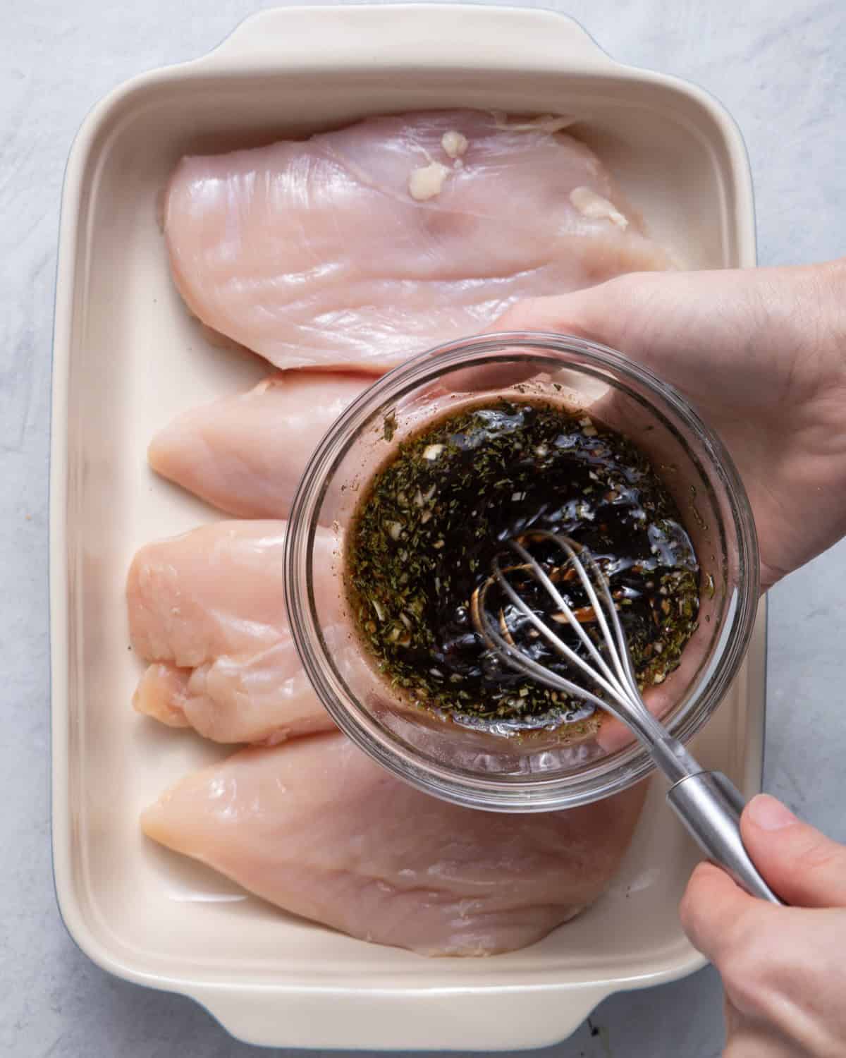 Hand holding small bowl with marinade on top of the chicken, whisking it.