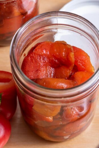 Roasted peppers in a jar with no lid, with another jar of roasted peppers and cut peppers nearby.