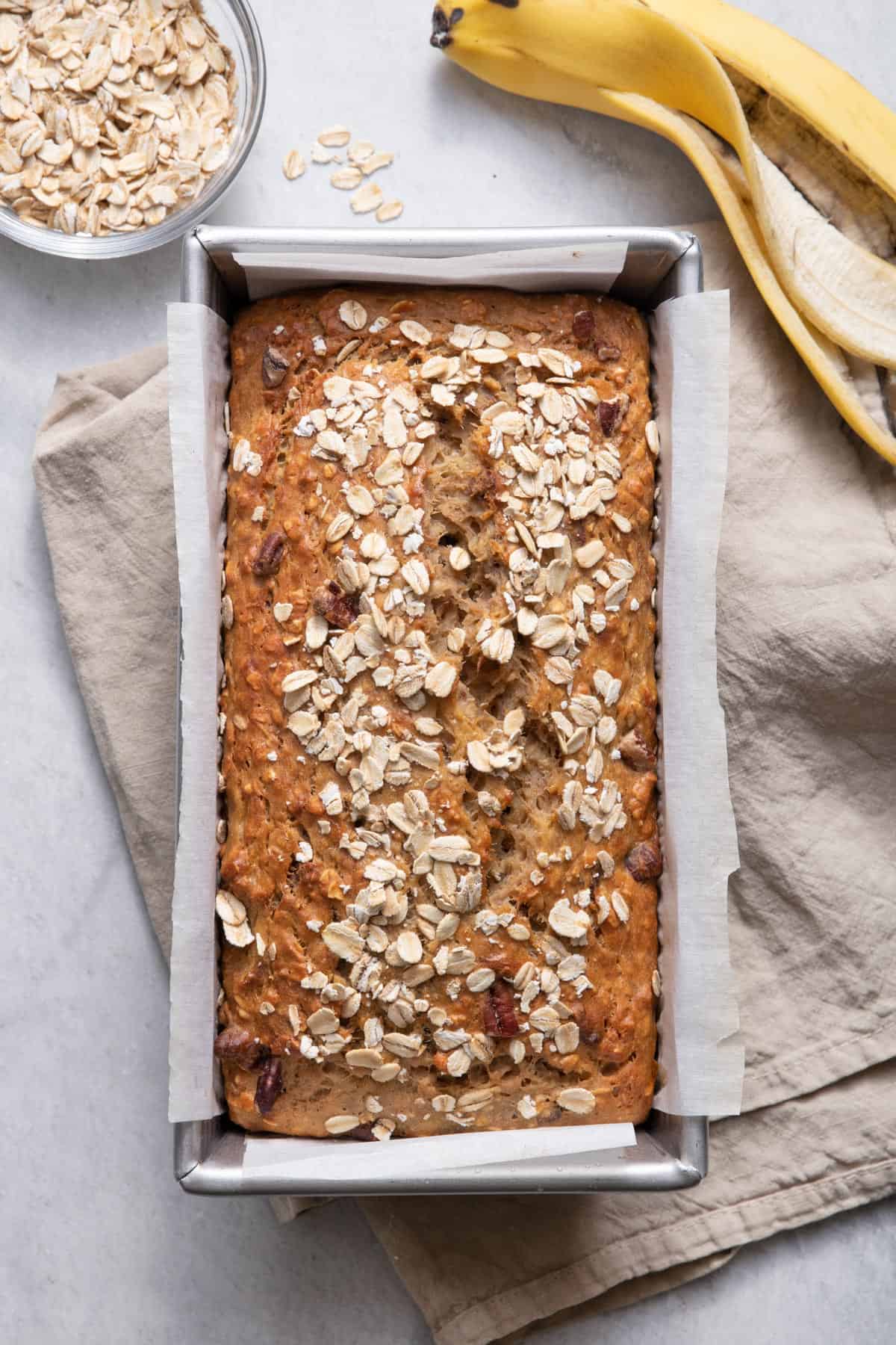 Oatmeal banana bread in loaf pan with oats nearby and banana peel