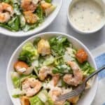 Two bowls of shrimp caesar salad with yogurt dressing on the side