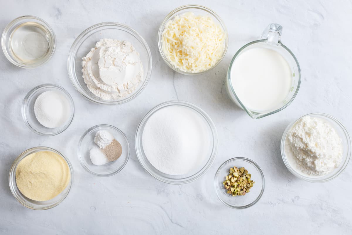 ingredients for recipe in individual bowls: simple syrup, sugar, semolina, baking bowder, instant yeast, salt, milk, crushed pistachios, ricotta, and mozzerella