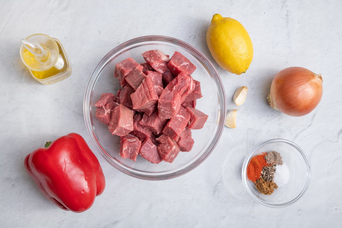 ingredients for grilled beef kabobs: diced beef, red pepper, lemon, onion, garlic cloves, olive oil, and seasonings