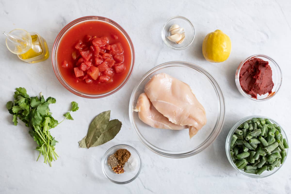 ingredients for recipe in individual bowls: olive oil in jar, diced canned tomatoes, fresh cilantro, bay leaves, spices, 2 chicken breast, garlic cloves, whole lemon, tomato paste, and frozen green beans