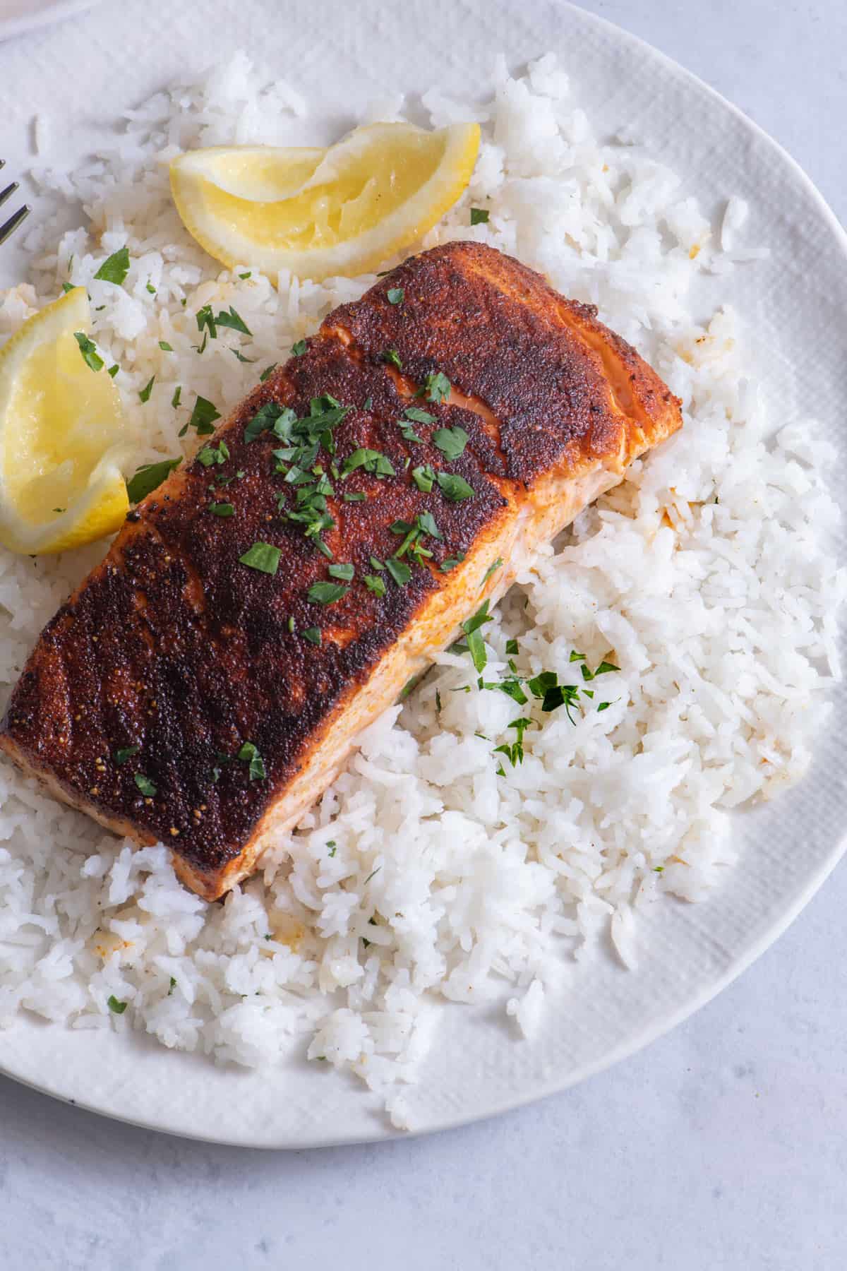 Large white serving plate full of basmati rice, topped with blackened salmon filet, and garnished with fresh parsley and two squeezed lemon slice.