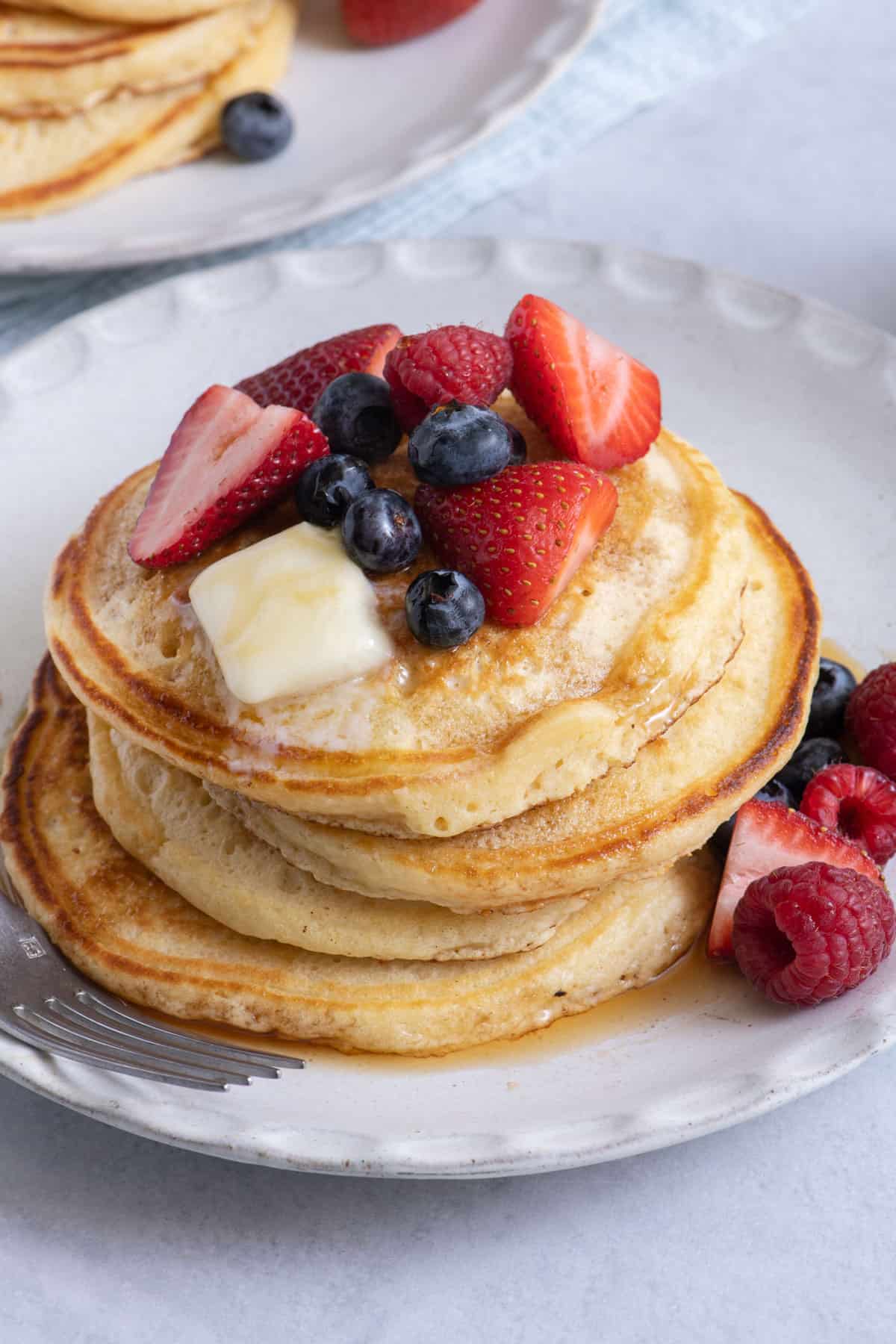 Stack of four small homemade pancakes topped with butter, strawberries, raspberries, and blueberries and served on a white plate with fork off to side.