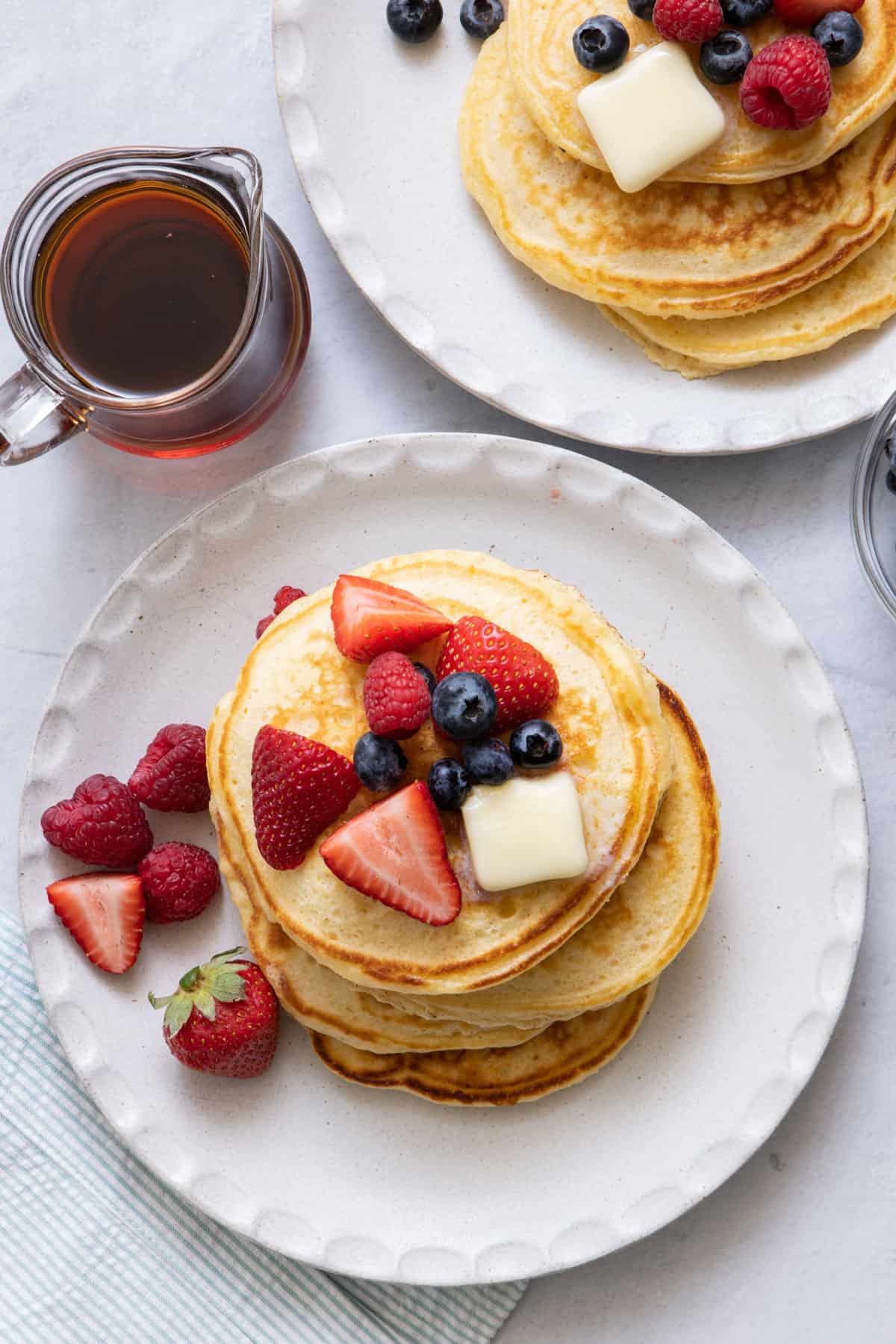 Overhead view of a stack of pancakes topped with fresh berries and butter on a large white plate with a side of maple syrup in glass carafe for pouring.