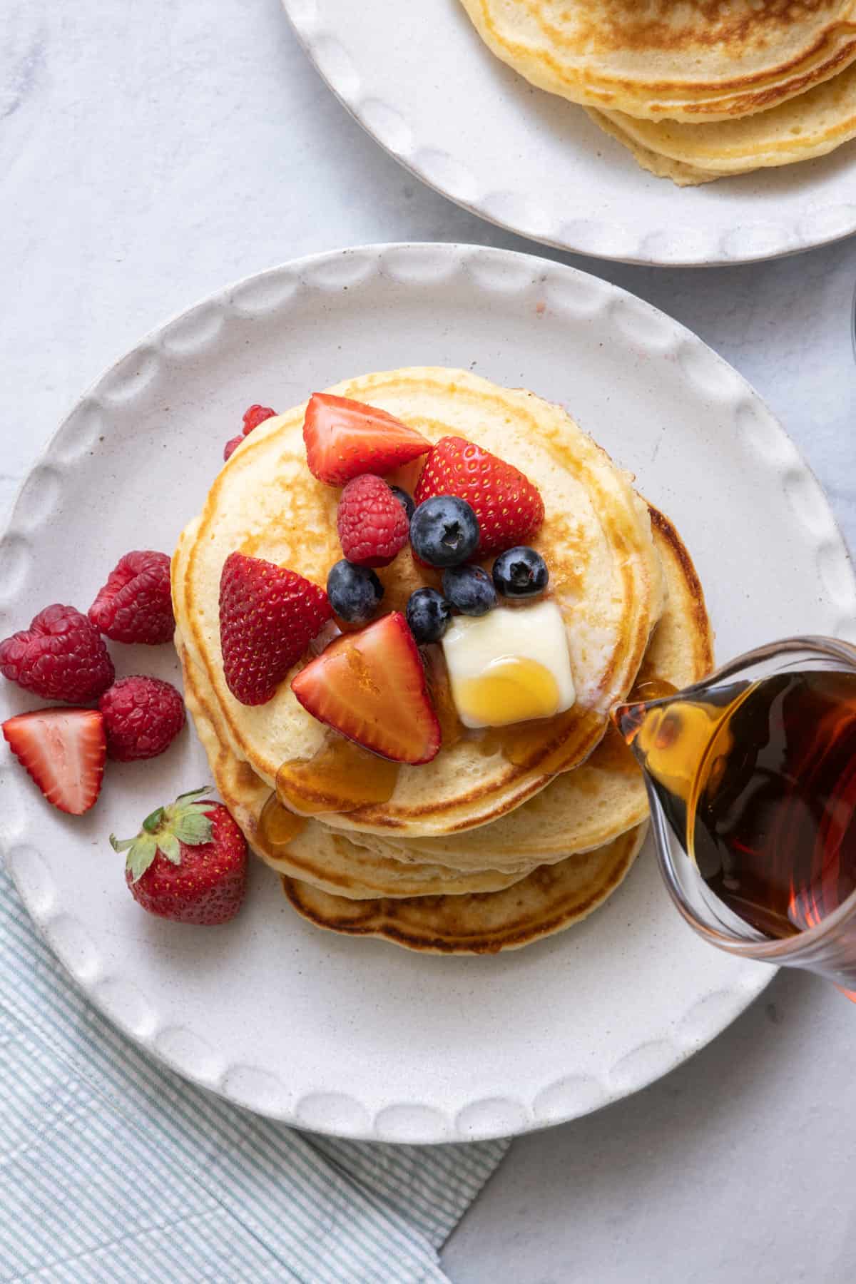 Maple syrup being poured over a stack of pancakes that is topped with fresh berries and butter.