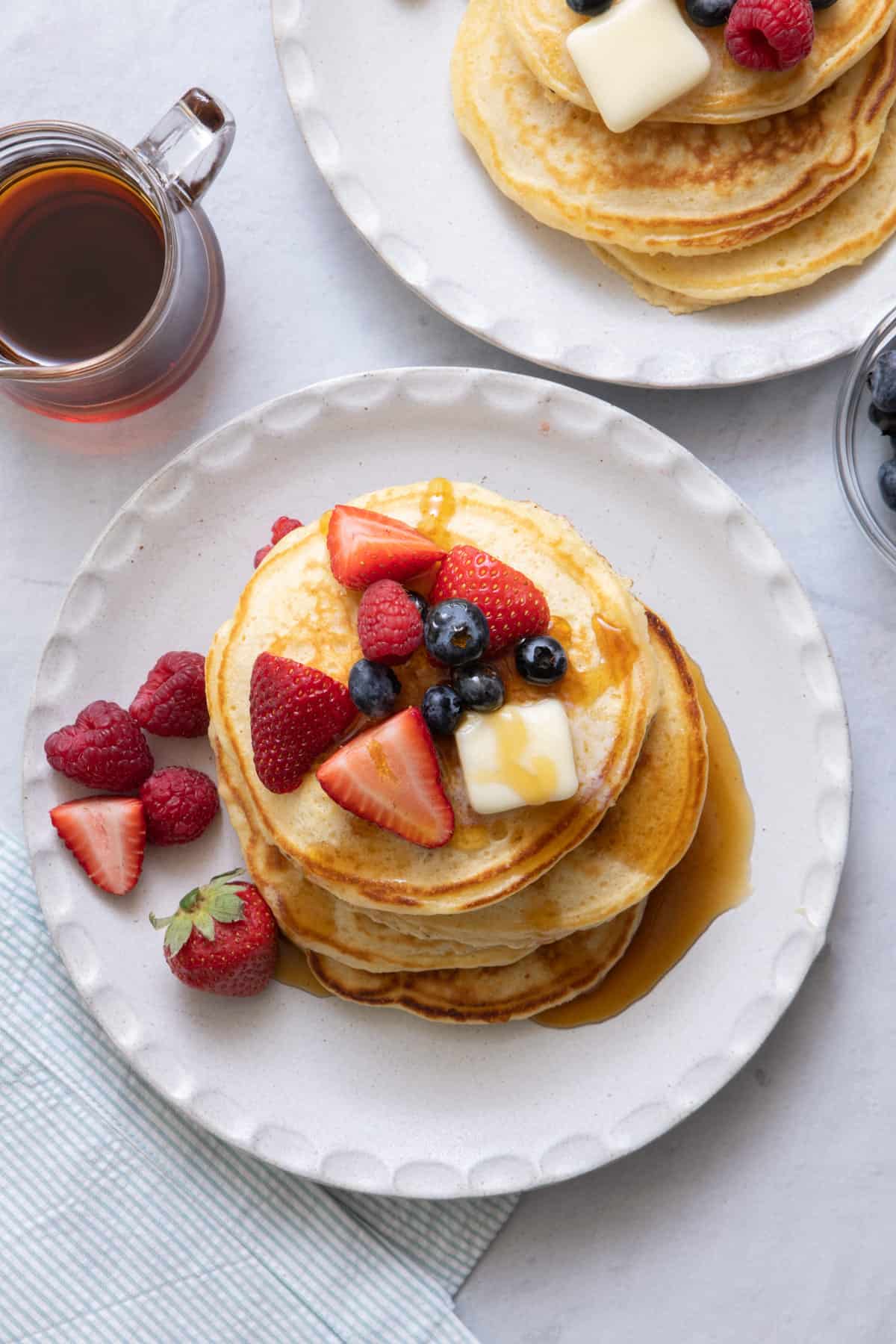 Stack of pancakes topped with maple syrup, berries, and butter on a large white plate.