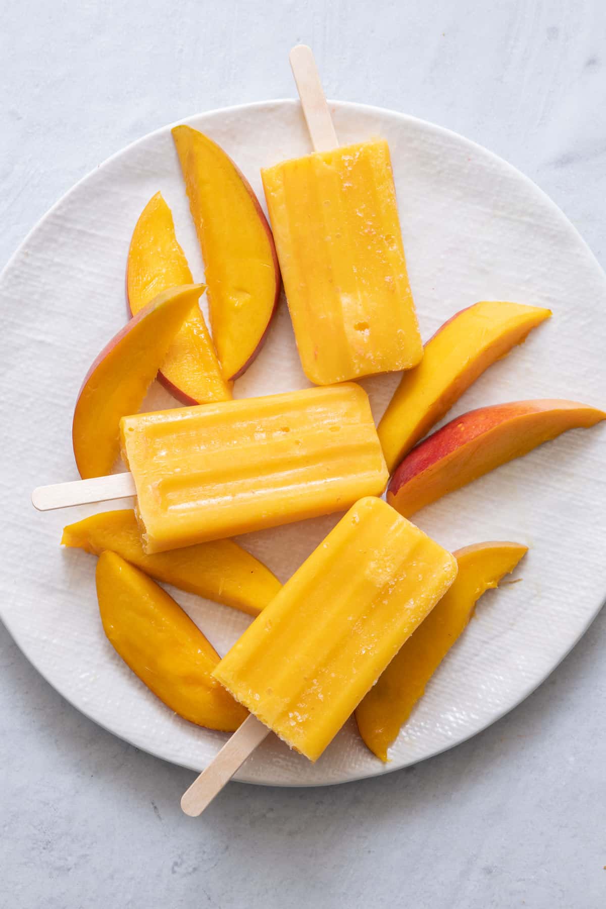 3 homemade mango popsicles on a large white plate with fresh cut mango.
