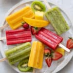 Homemade Mango, Kiwi, and Strawberry popsicles on a large round platter with ice cubes and fresh cut fruit.