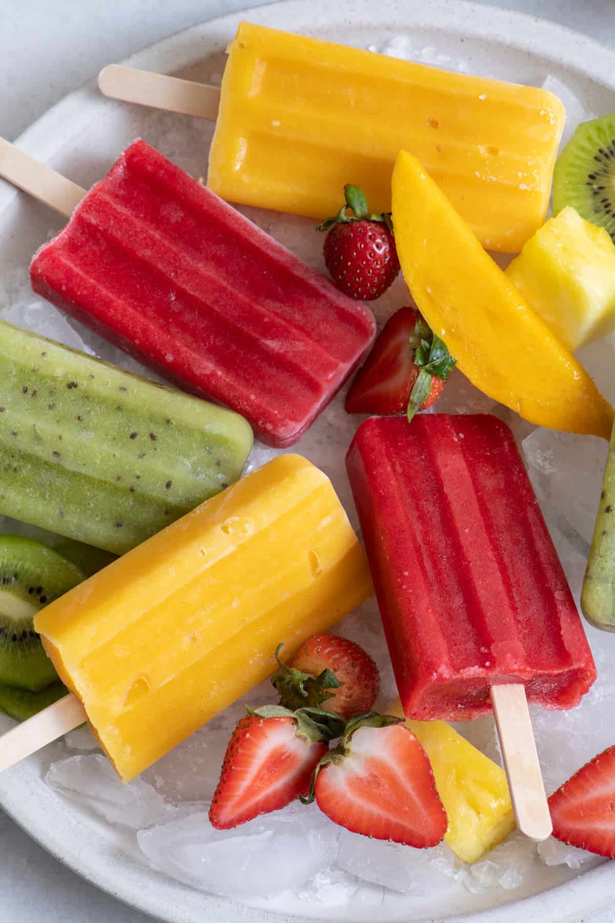 Homemade Mango, Kiwi, and Strawberry popsicles on a large round platter with ice cubes nd fresh cut fruit.