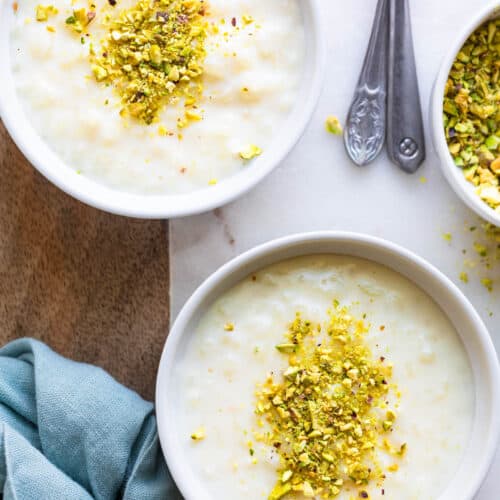 Overhead shot of two bowls of Lebanese Rice Pudding garnished with chopped pistachios and two serving spoons set off to side.