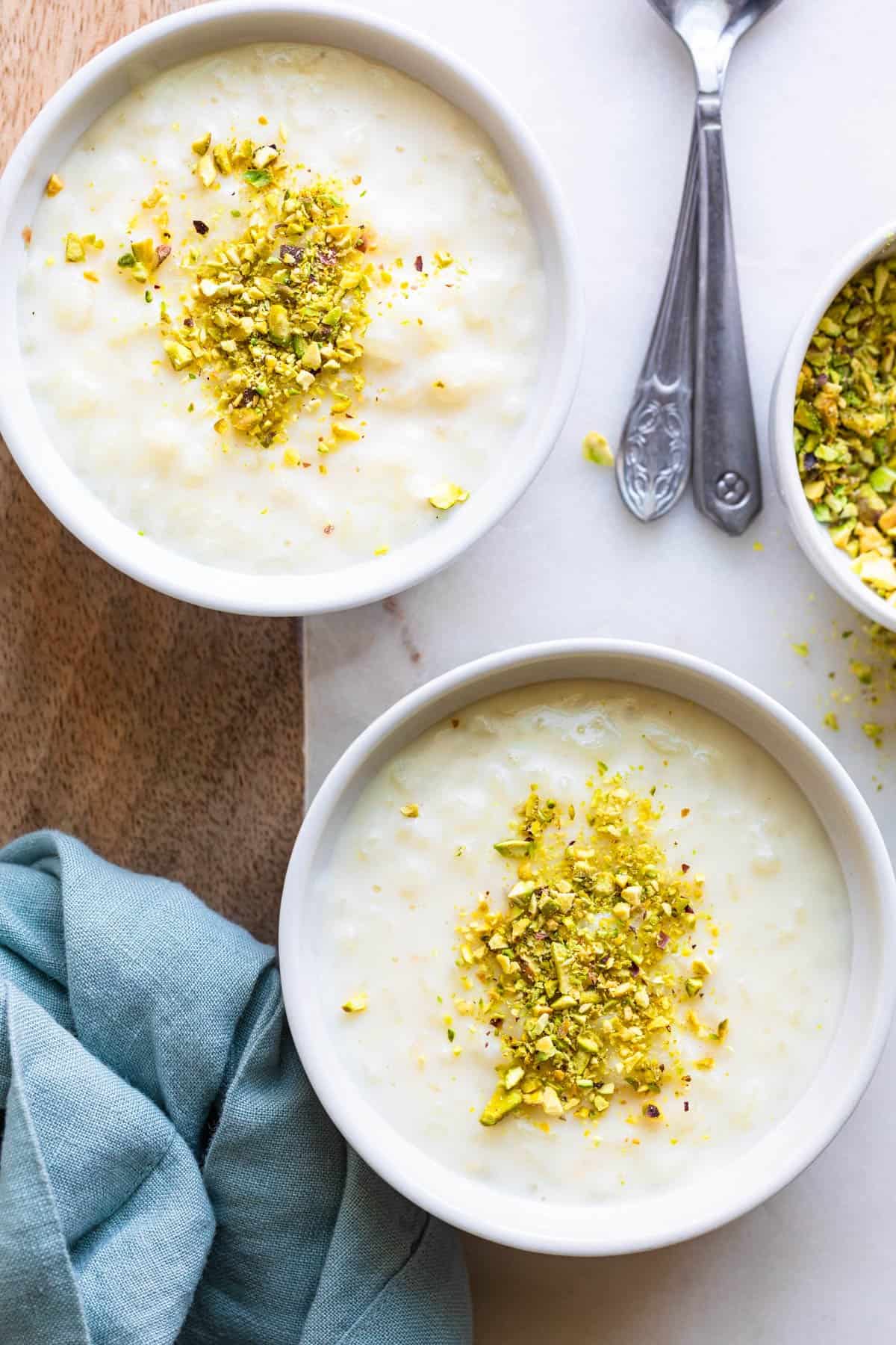 Overhead shot of two bowls of Lebanese Rice Pudding garnished with chopped pistachios and two serving spoons set off to side.