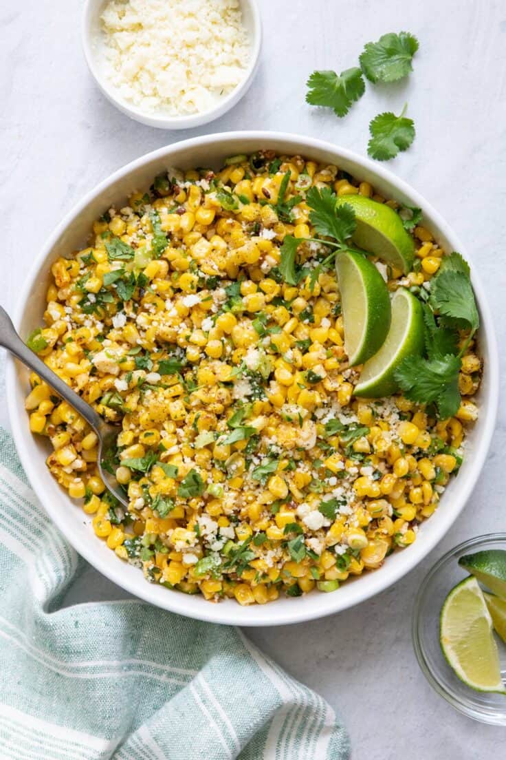 Overhead shot of Mexican Street Corn Salad in large white bowl with lime wedges and side of cotija cheese crumbles.