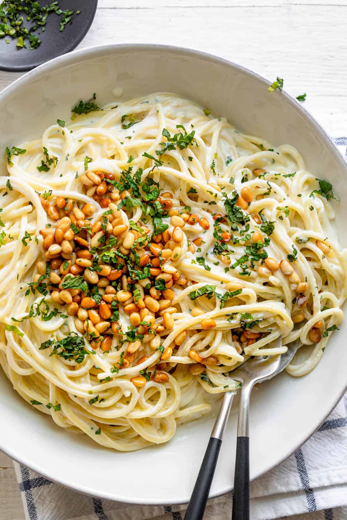 Serving dish of spaghetti noodles in white sauce with herbs and pine nuts on top and serving spoon and fork tucked in noodles.
