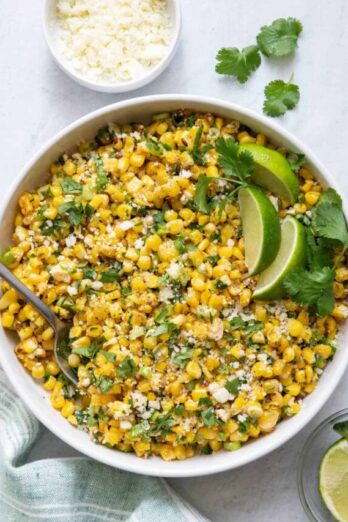 Overhead shot of Mexican Street Corn Salad in large white bowl with lime wedges and side of cotija cheese crumbles.