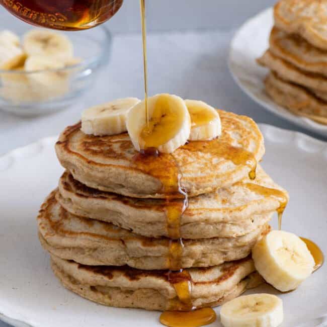 Maple syrup being poured over a stack of banana pancakes with fresh slices of bananas on top.