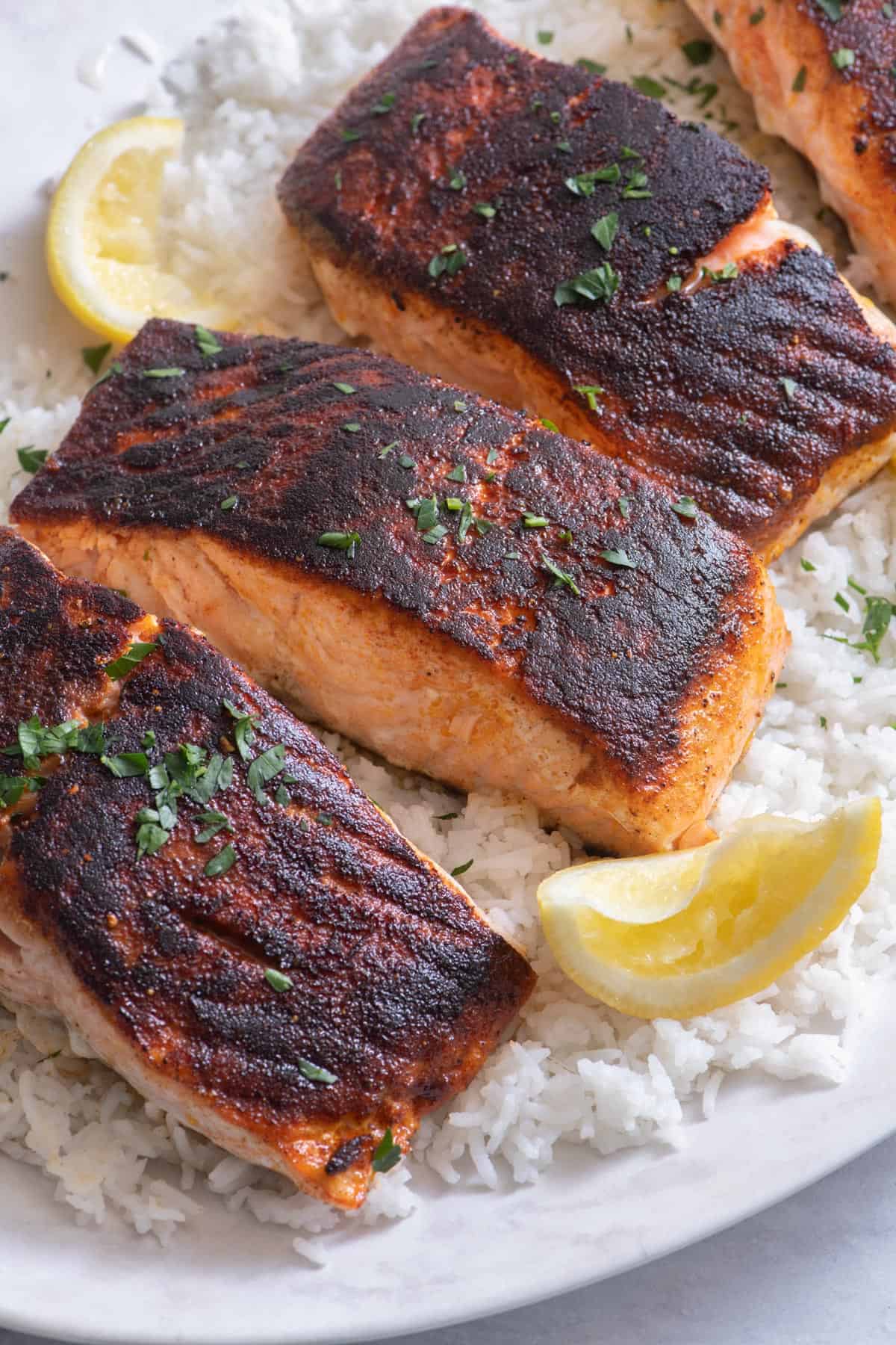 Close up of blackened salmon filets atop a bed of white rice and garnished with lemon wedges and parsley.