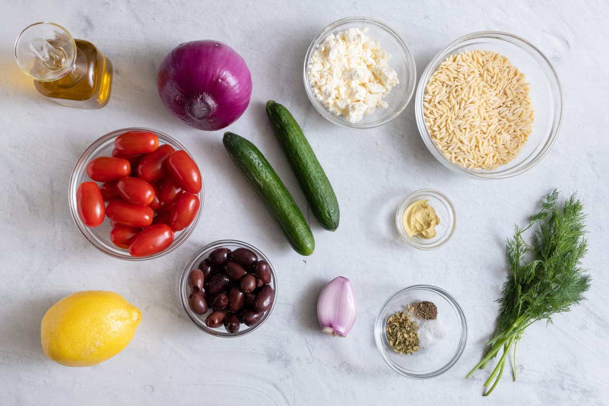 Ingredients for recipe before being prepped: olive oil, grape tomatoes, whole lemon, whole red onion, 2 Persian cucumbers, crumbled feta, uncooked orzo, garlic clove, spices, dijon mustard, and fresh dill.