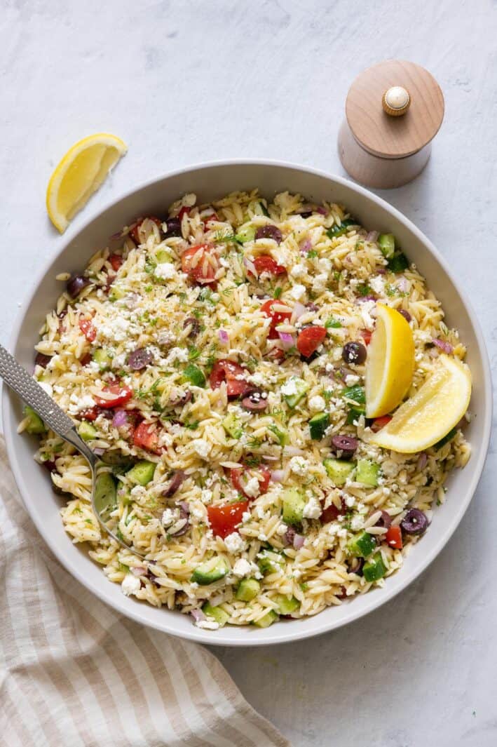 Large bowl of orzo salad with spoon dipped in and garnished with lemon wedges.