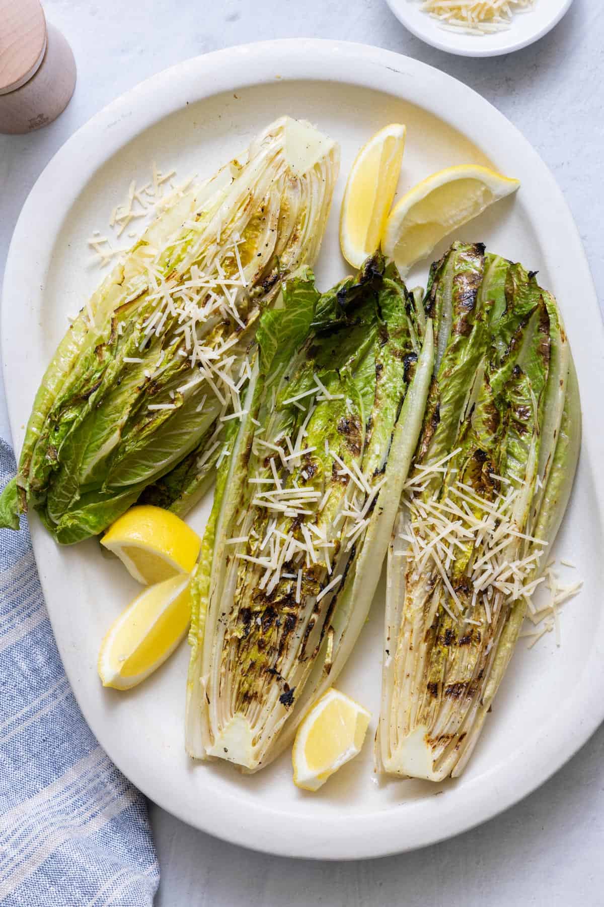 Large white oval platter with 3 halved grilled romaine lettuces sprinkled with shredded parmesan and garnished with lemon wedges.
