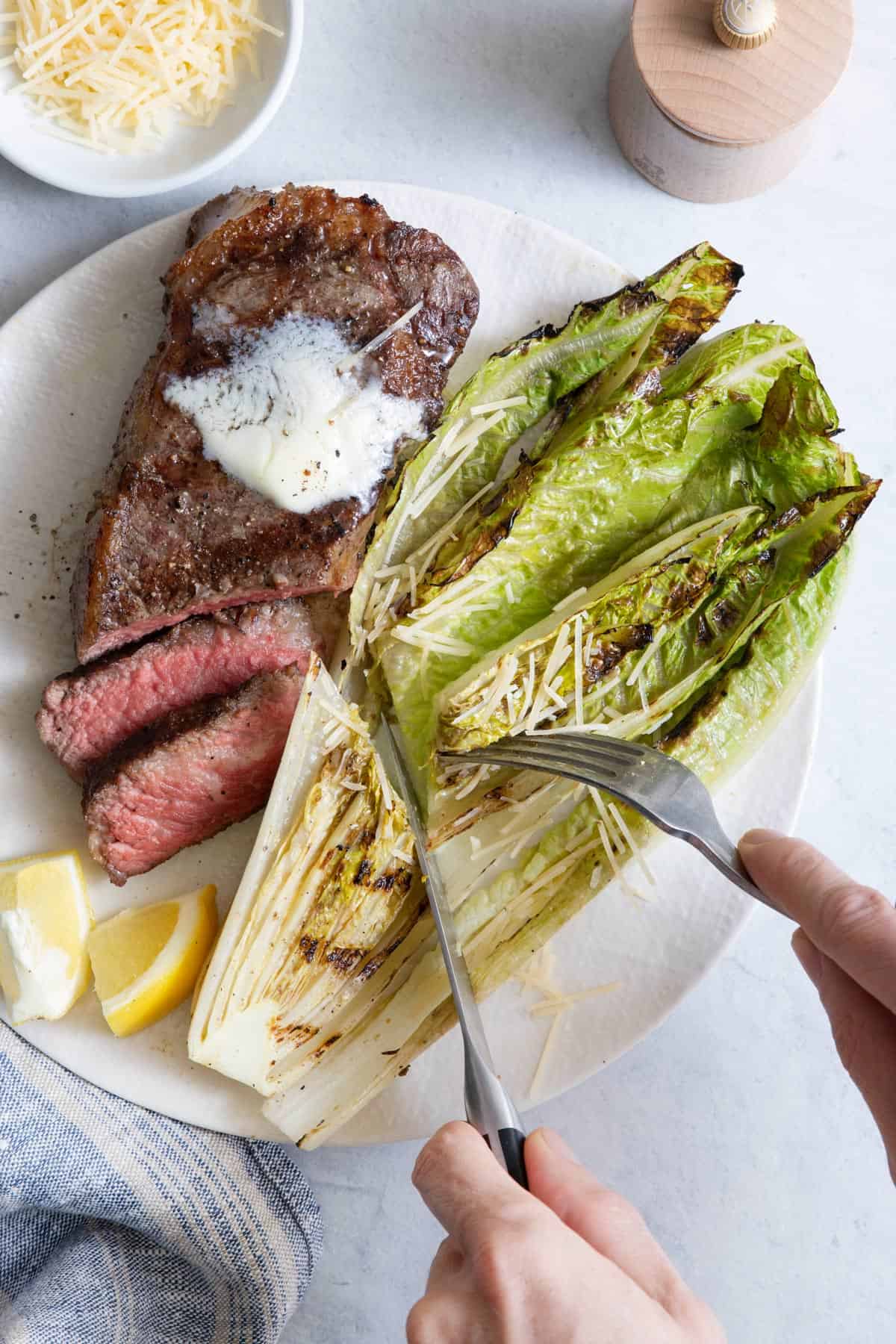 Fork and knife cutting romaine lettuce in half on large white plate with that's being served with a grilled steak.