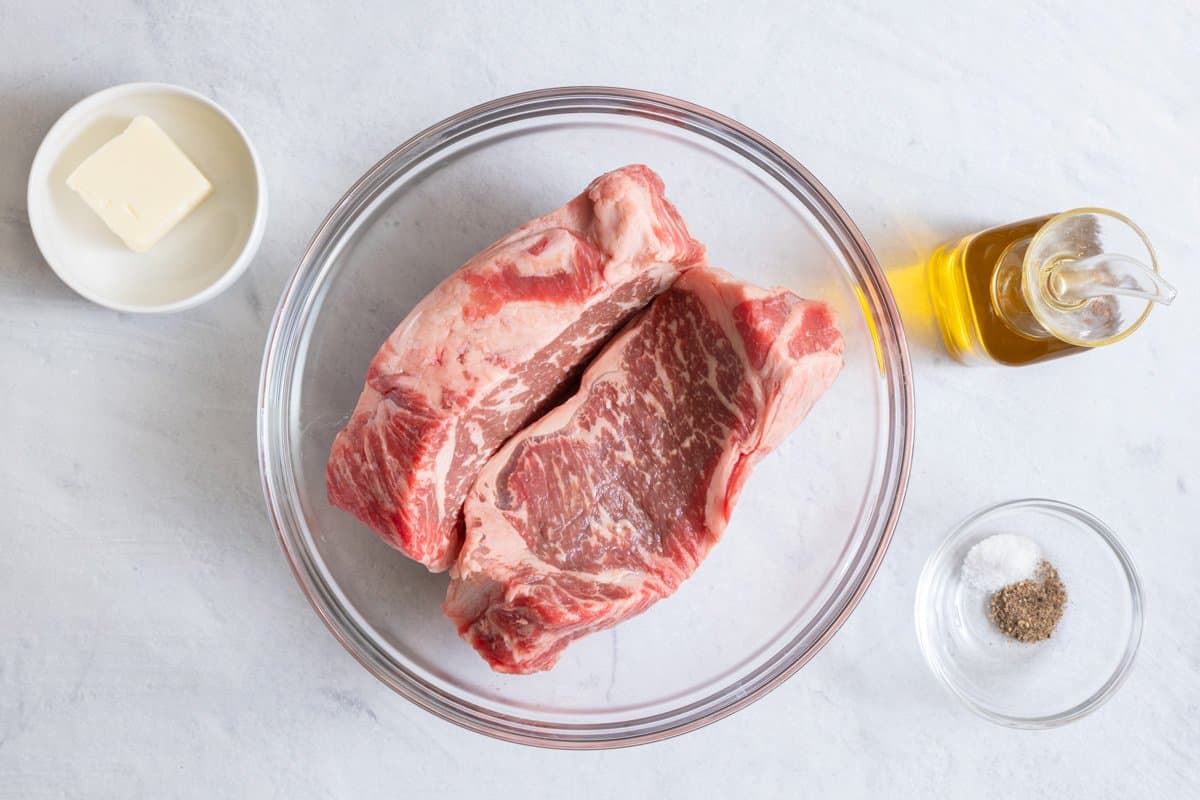 Ingredients for grill recipe: two steaks in a glass bowl, cube of butter, olive oil, salt, and pepper.