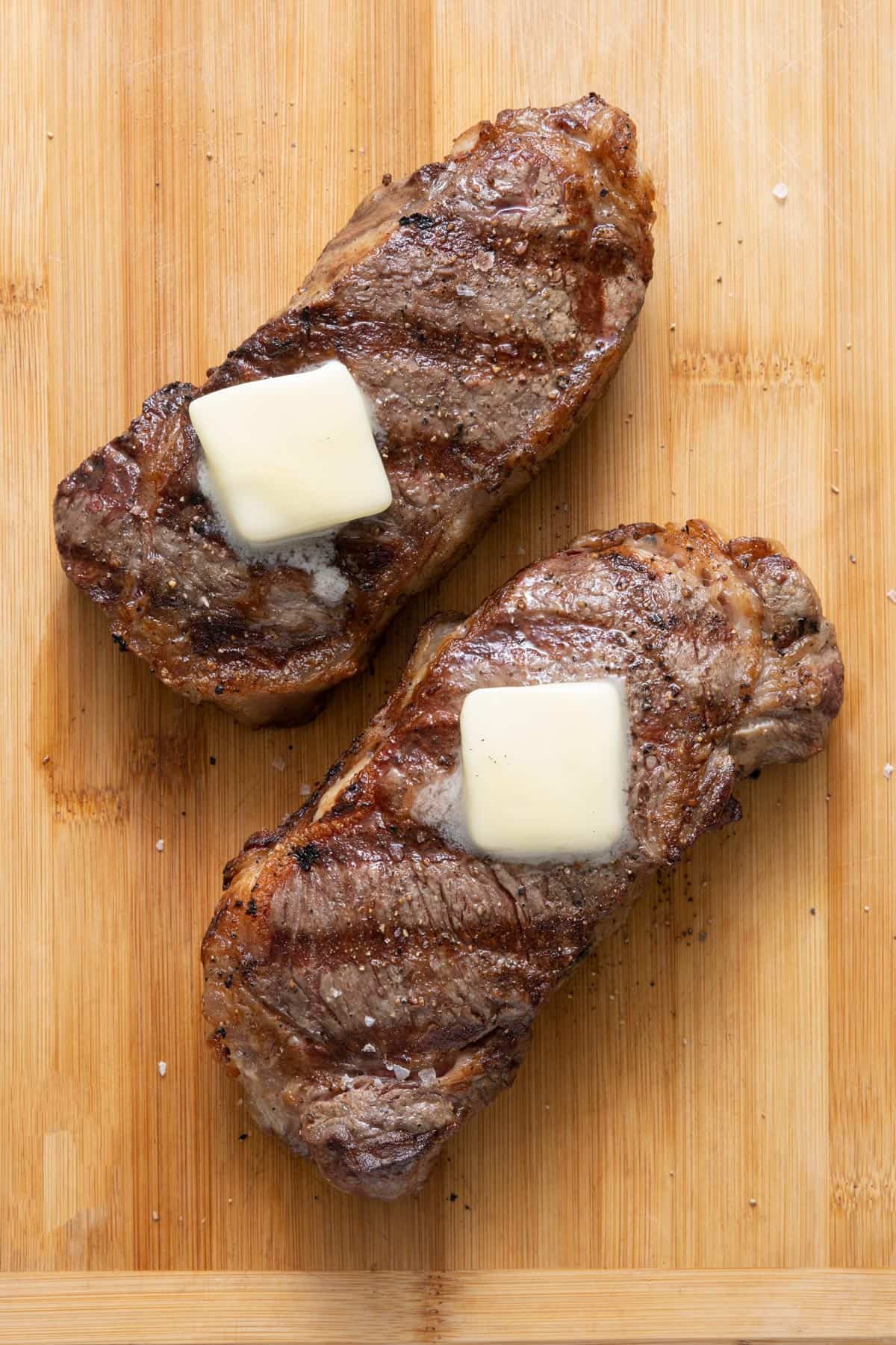 Two grilled steaks on cutting board with butter on top melting.