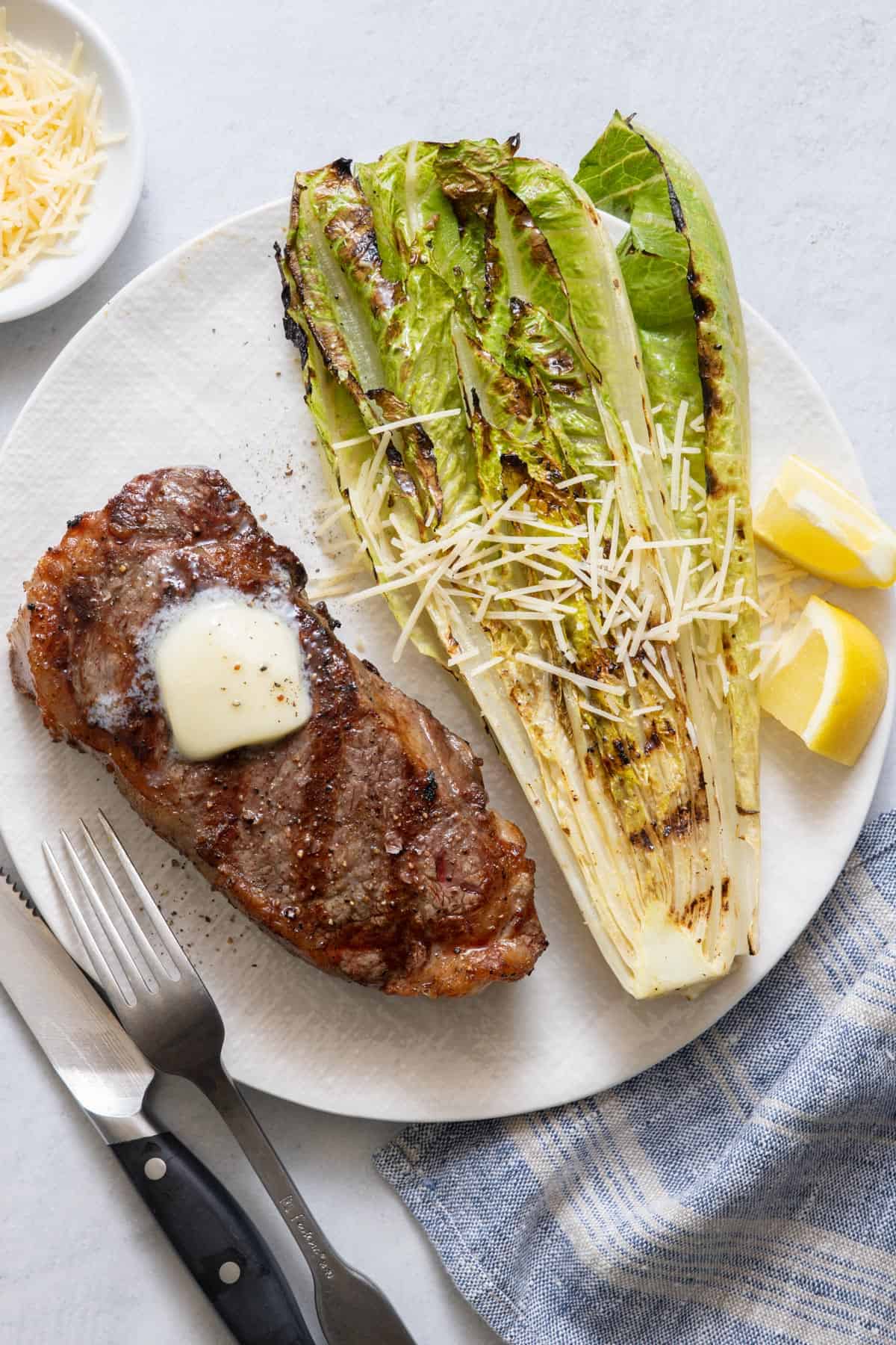 Grilled steak topped with melted butter on a large white plate with grilled romaine lettuce, lemon wedges, parmesan, and fork.