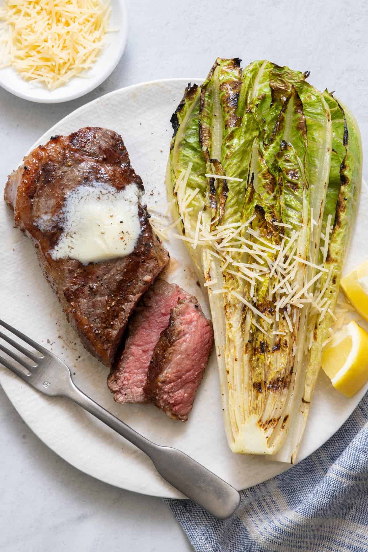 Grilled steak with two slices cut from it topped with melted butter on a large white plate with grilled romaine lettuce, lemon wedges, parmesan, and fork.