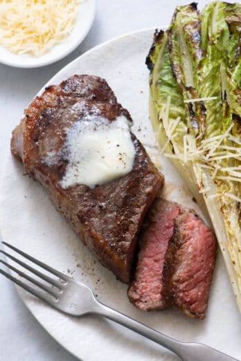 Close up of grilled steak with two slices cut from it topped with melted butter on a large white plate with grilled romaine lettuce, parmesan, and fork.
