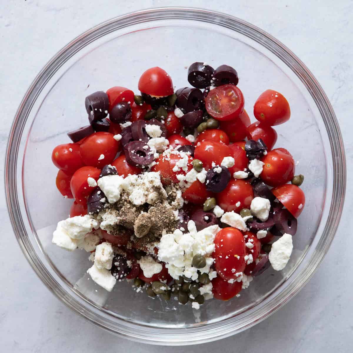 Tomatoes, olives, feta cheese, capers and black pepper in large bowl before mixed.