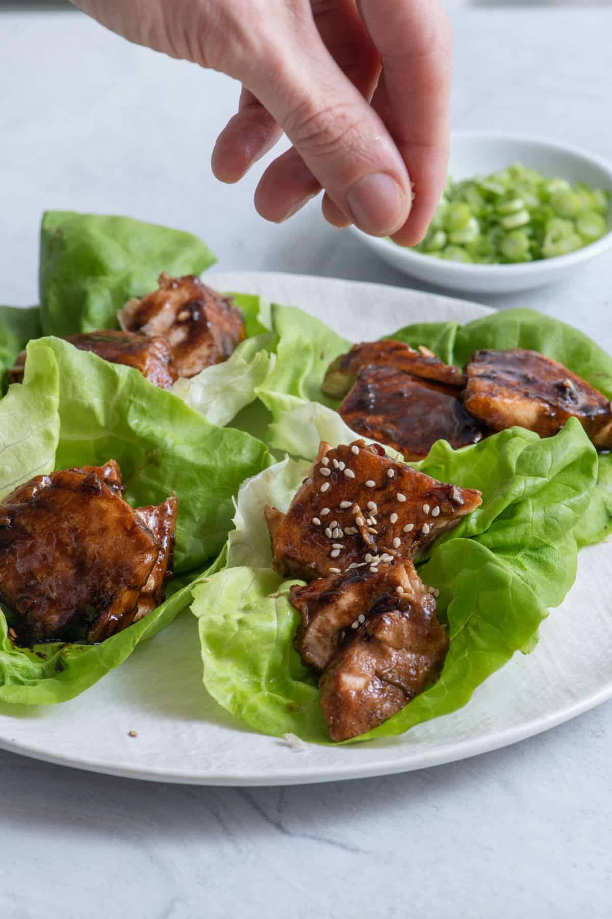 Four pieces of Bibb lettuce each with chunks of salmon and a hand garnishing with sesame seeds.