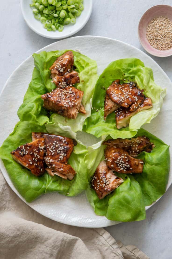 Four Salmon Lettuce Wraps on large white plate with a side dish of both scallions and sesame seeds.