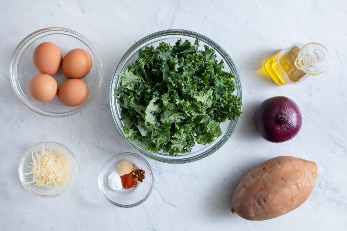 Ingredients for recipe: four uncooked shelled eggs, Parmesan cheese, spices, chopped kale, sweet potato, red onion, and oil.