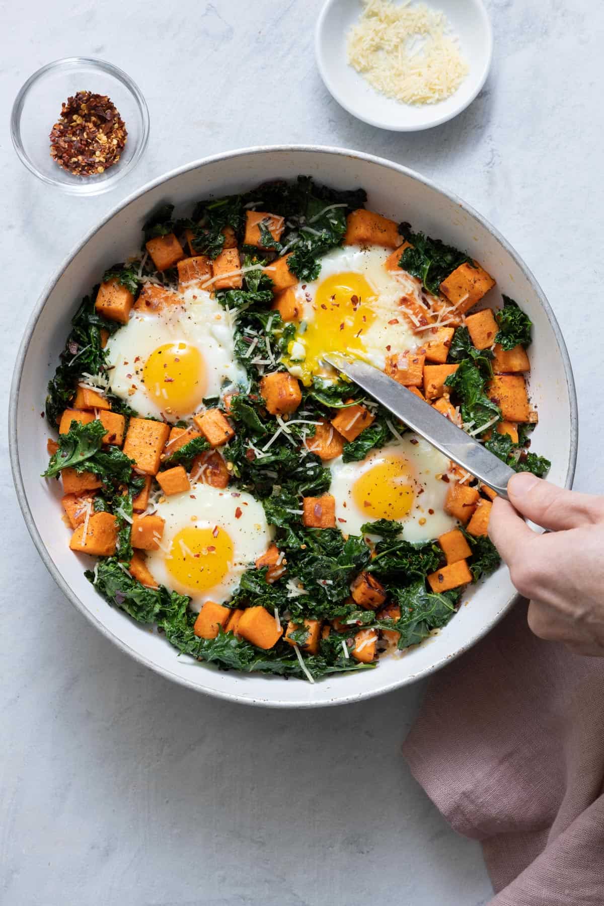 Skillet with the cooked eggs and garnished with parmesan cheese and red pepper flakes and knife cutting one of the yolks showing soft yolk spilling into kale/sweet potatoes.