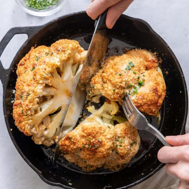 Hands with knife and fork cutting roasted cauliflower in cast iron skillet.