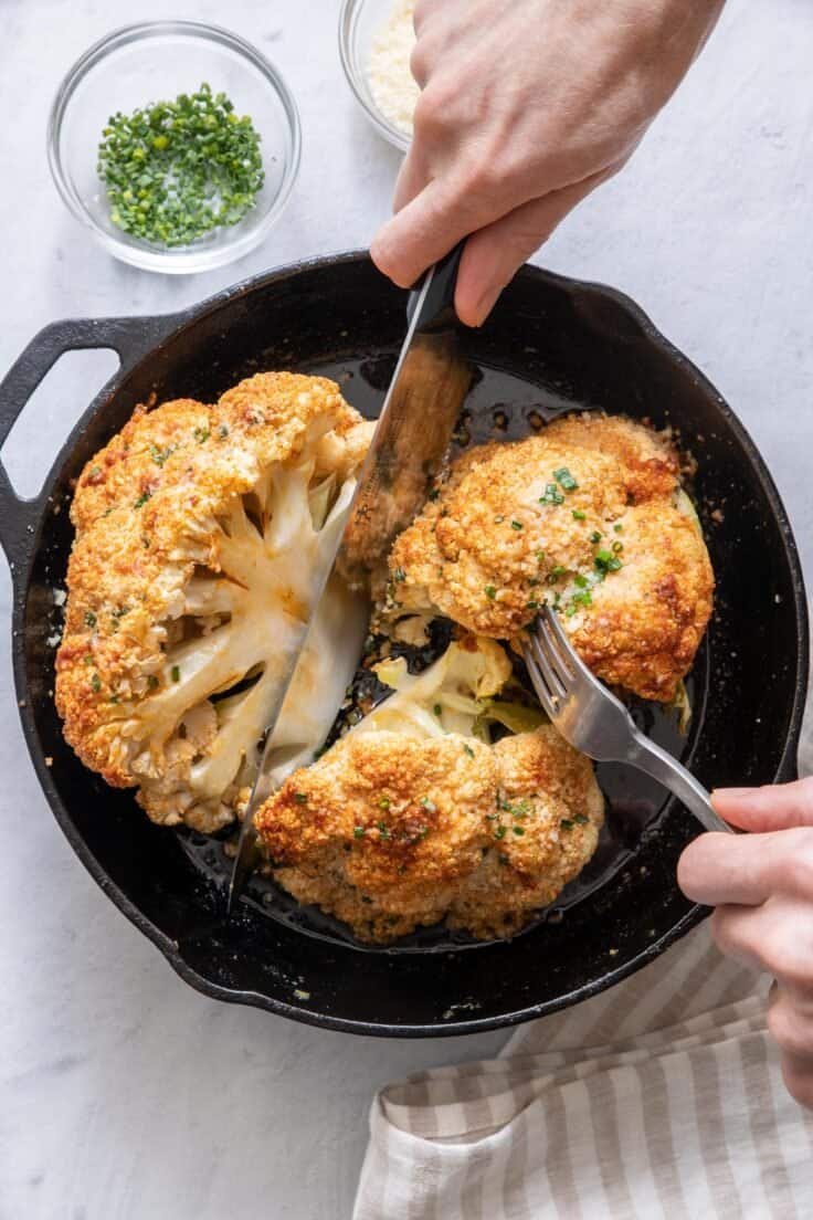 Hands with knife and fork cutting roasted cauliflower in cast iron skillet.
