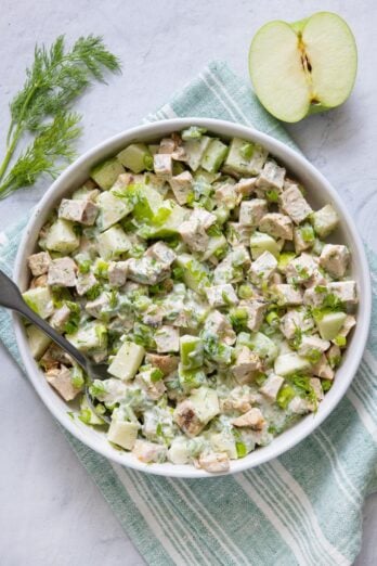 Yogurt chicken salad in a large white serving bowl garnished with fresh dill and green onions and half an apple off to side.