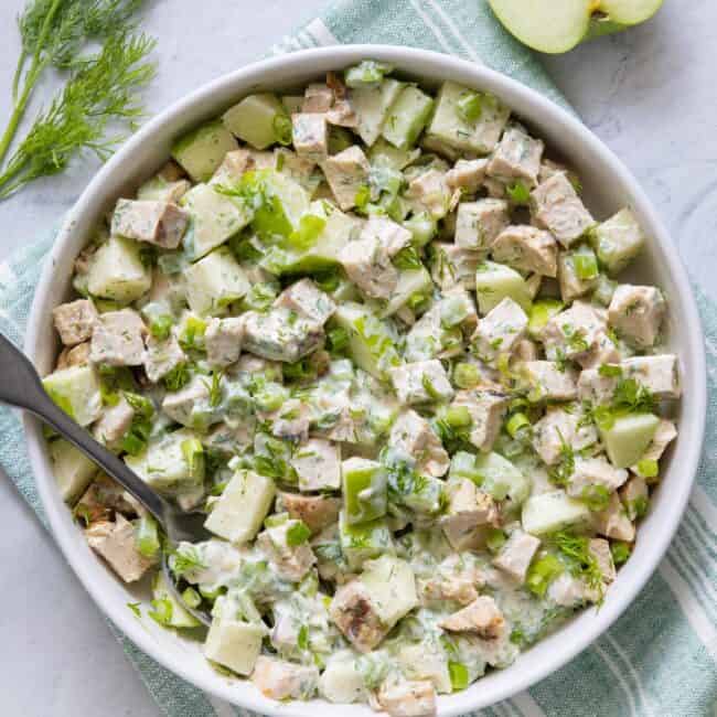 Yogurt chicken salad in a large white serving bowl garnished with fresh dill and green onions and half an apple off to side.