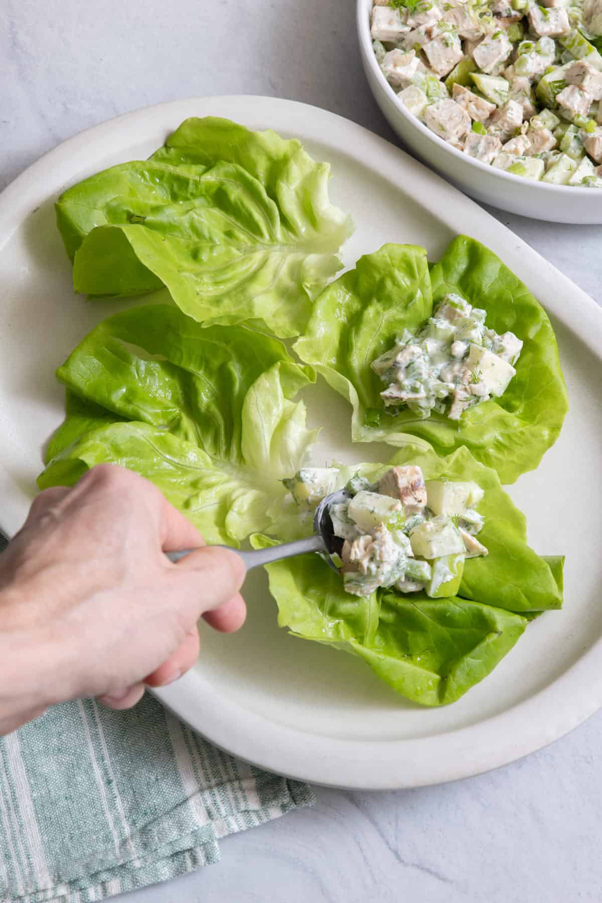 Large platter with four butter lettuce cups with a hand filling them with chicken salad recipe.