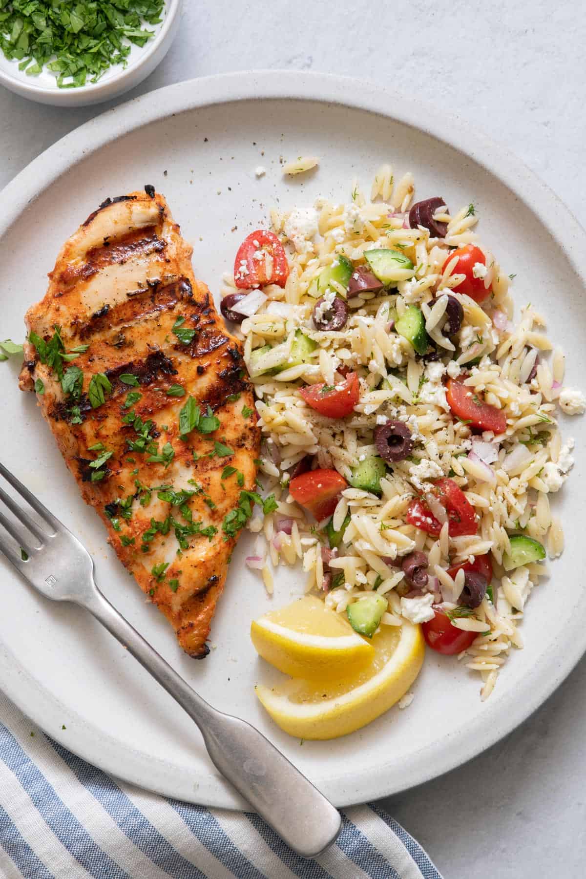 Plate with grilled chicken breast and next to orzo salad and garnished with fresh parsley and lemon wedges.