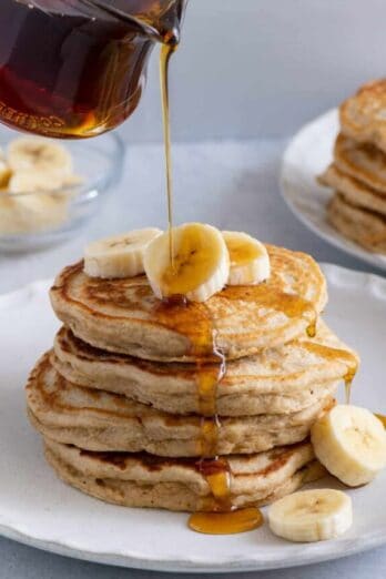 Maple syrup being poured over a stack of banana pancakes with fresh slices of bananas on top.