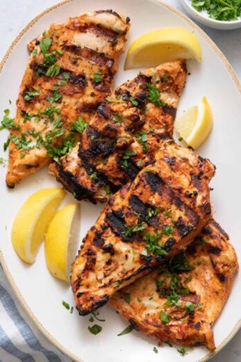 Over head shot showing 4 grilled chicken breasts on plate garnished with lemon wedges and parsley.