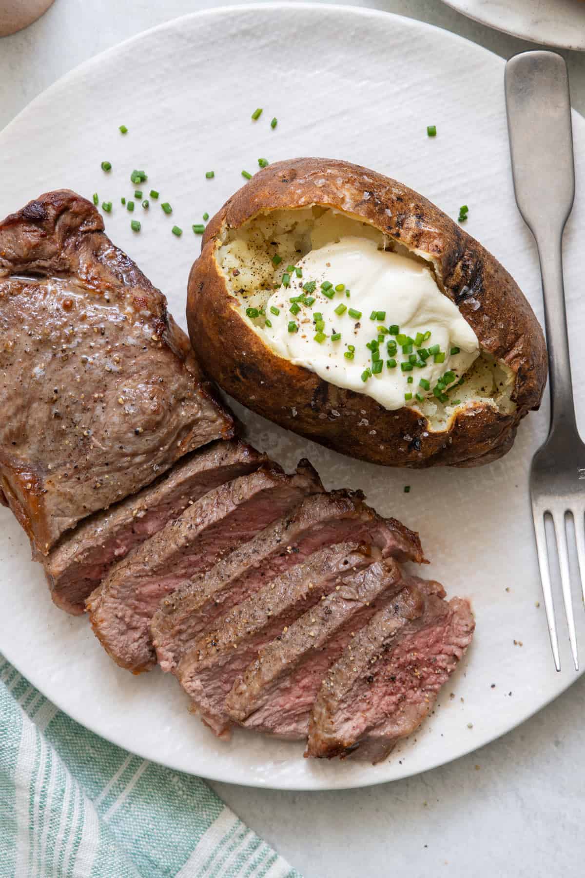 Air fryer baked potato topped with butter and chives on plate with a steak filet sliced halfway.