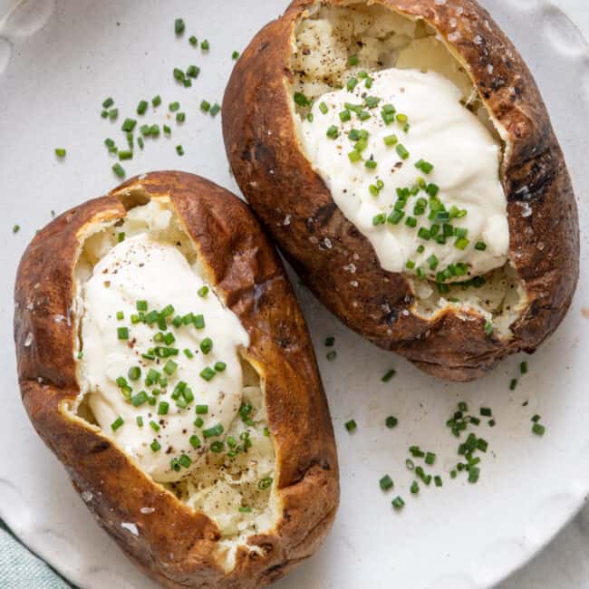 Two baked potatoes on plate topped with sour cream and chopped chives.