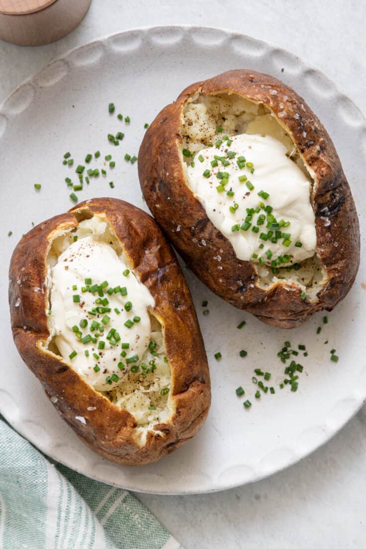 Two baked potatoes on plate topped with sour cream and chopped chives.