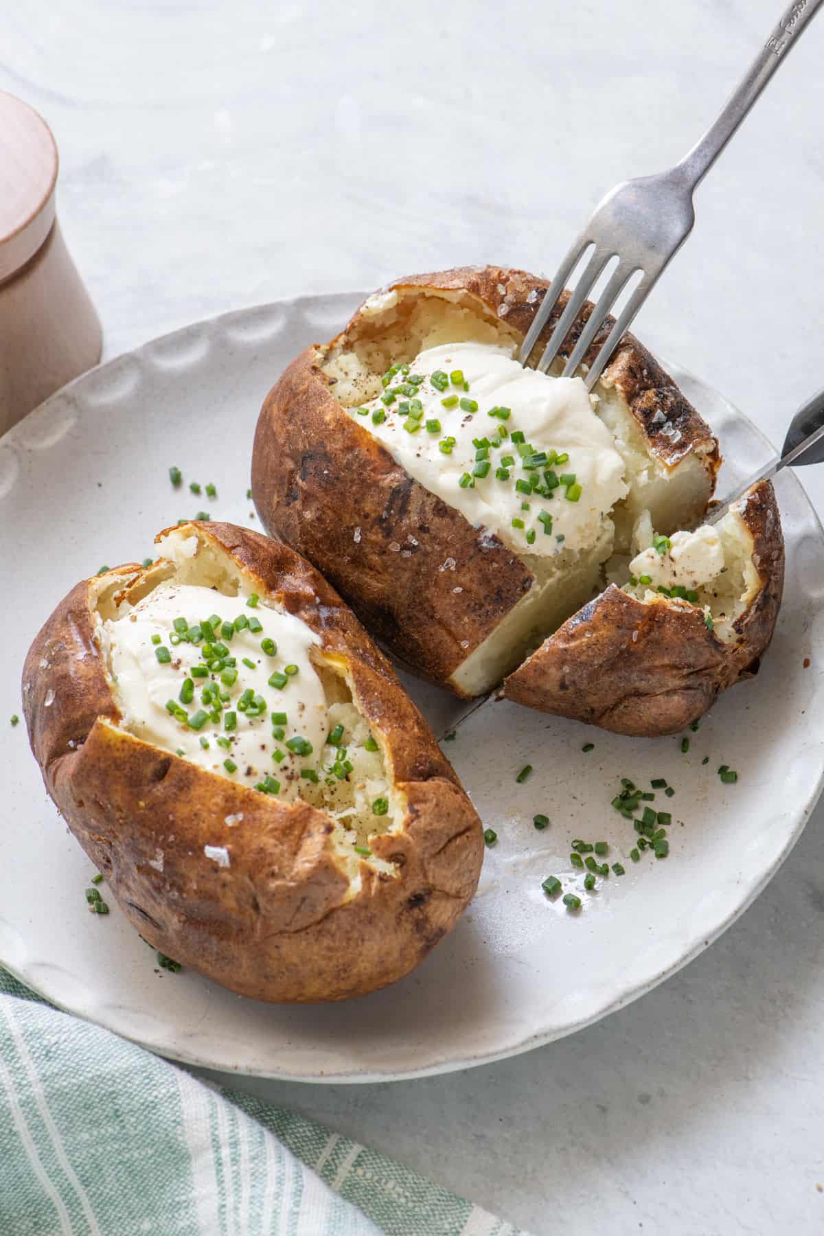 Two baked potatoes on plate topped with sour cream and chopped chives, cutting into one of them with knife and fork.