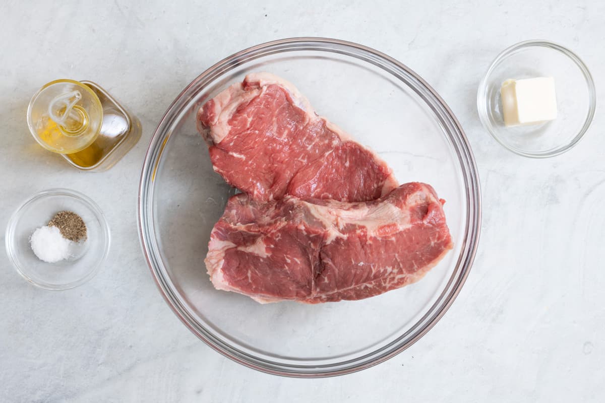 Ingredients for air fryer recipe: oil, salt, pepper, 2 steaks, and butter in individual bowls.